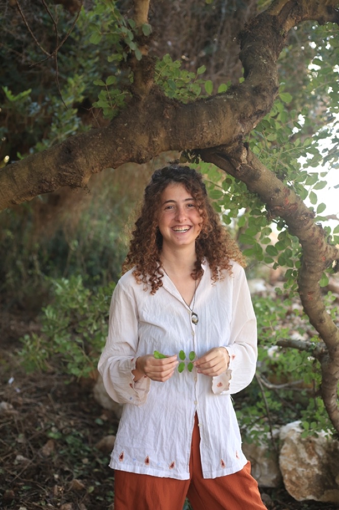 קארין מחייכת מתחת לעץ, מחזיקה עלה
